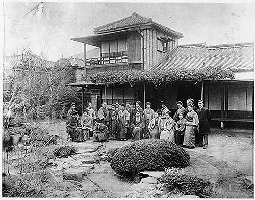 Eine Außenansicht der Tsuda Universität kurz nach ihrer Gründung. Vor ihr stehen und sitzen Dozent*innen und Studentinnen der Universität in Kimonos oder vormellem Anzug. Es sind etwa 15 Personen zu sehen, hinter ihnen das Universitätsgebäude mit etwa zwei Stockwerken. Um sie herum ist ein Garten.